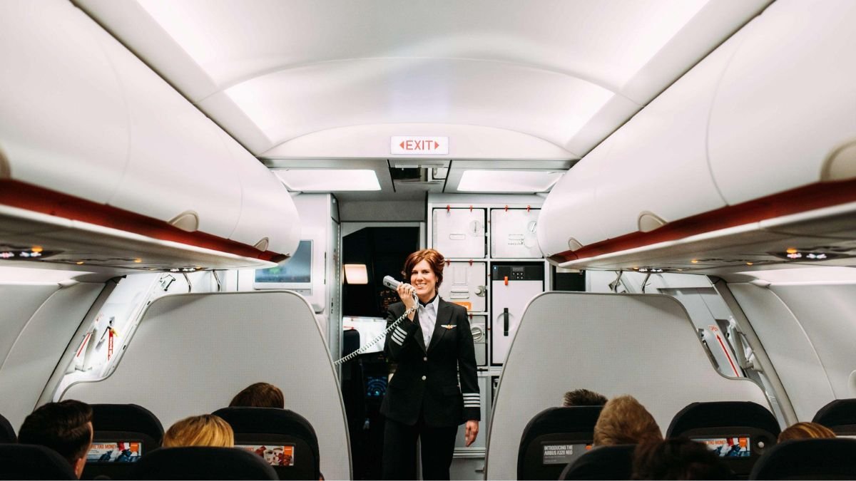 Comissário de voo realizando anúncio PA em inglês dentro da cabine de passageiros de uma aeronave